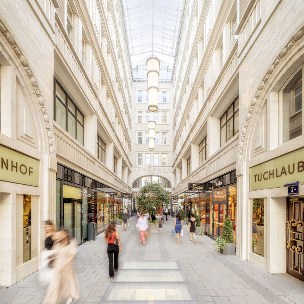 Bright covered shopping arcade with glass roof and people walking between boutique storefronts.